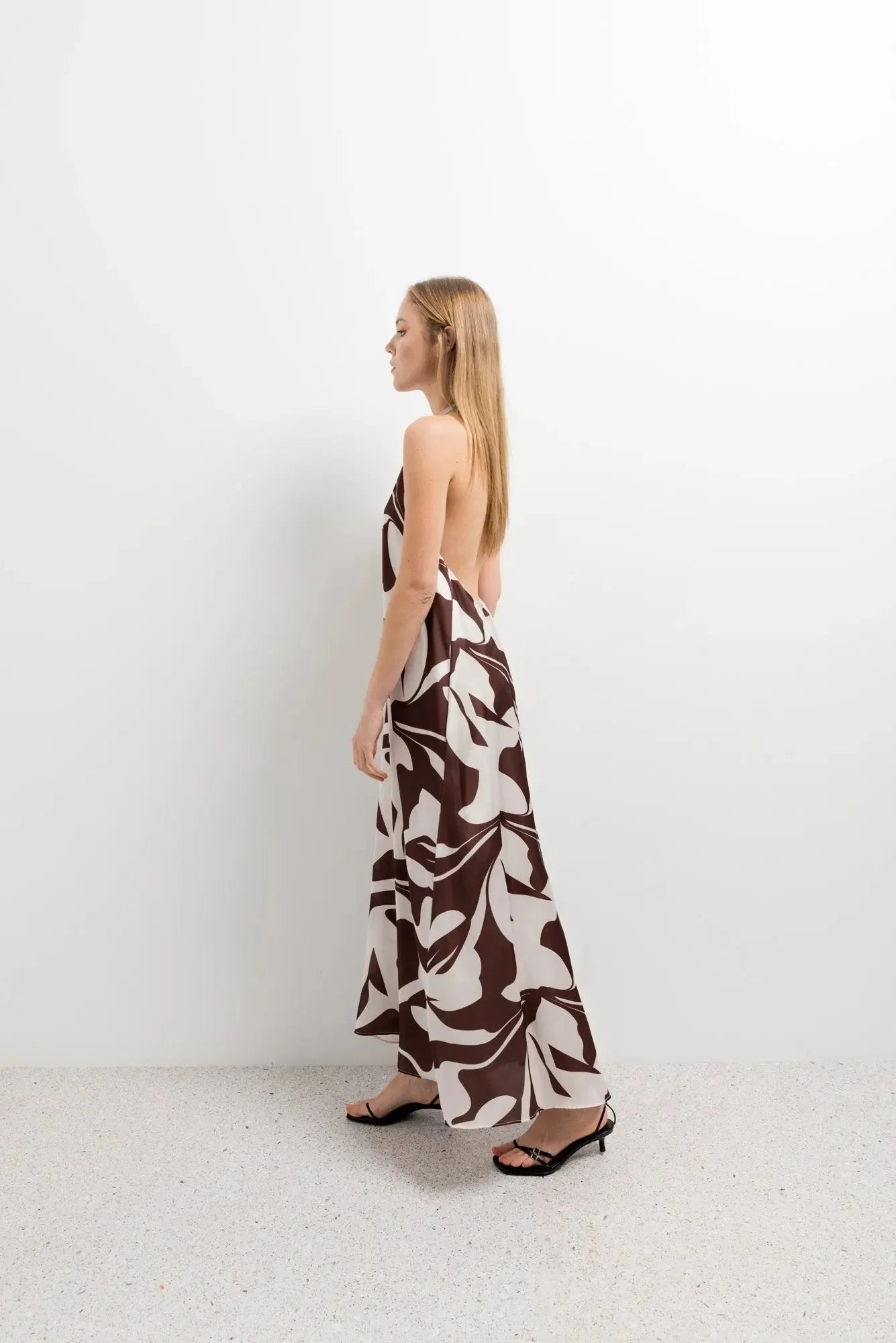 Robe Longue Bohême Imprimée Chocolat & Blanc Dos Nu Fluide avec Col Licou, femme de profil dans un décor minimaliste, mettant en avant le design élégant.