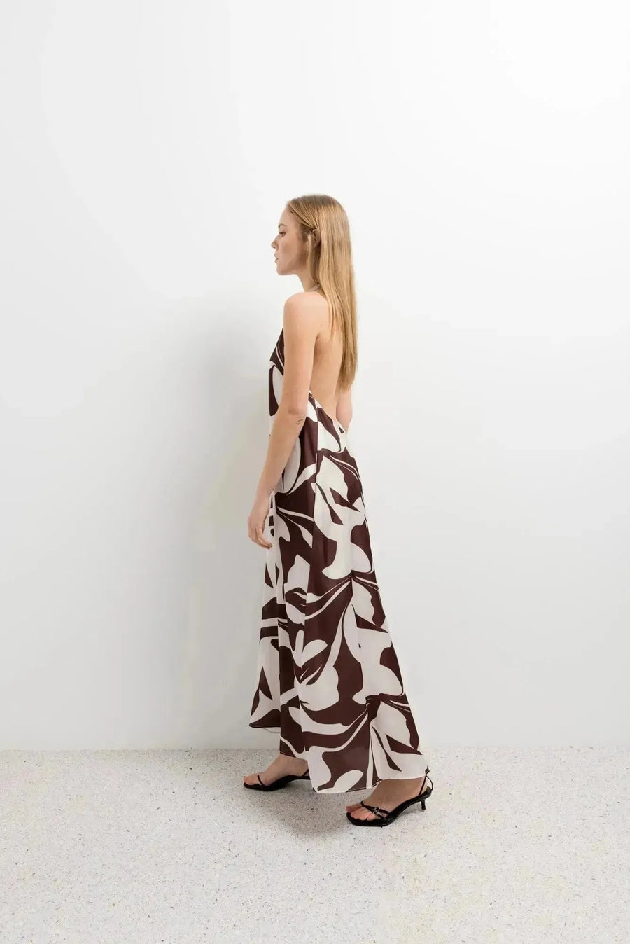 Robe Longue Bohême Imprimée Chocolat & Blanc Dos Nu Fluide avec Col Licou, femme de profil dans un décor minimaliste, mettant en avant le design élégant.