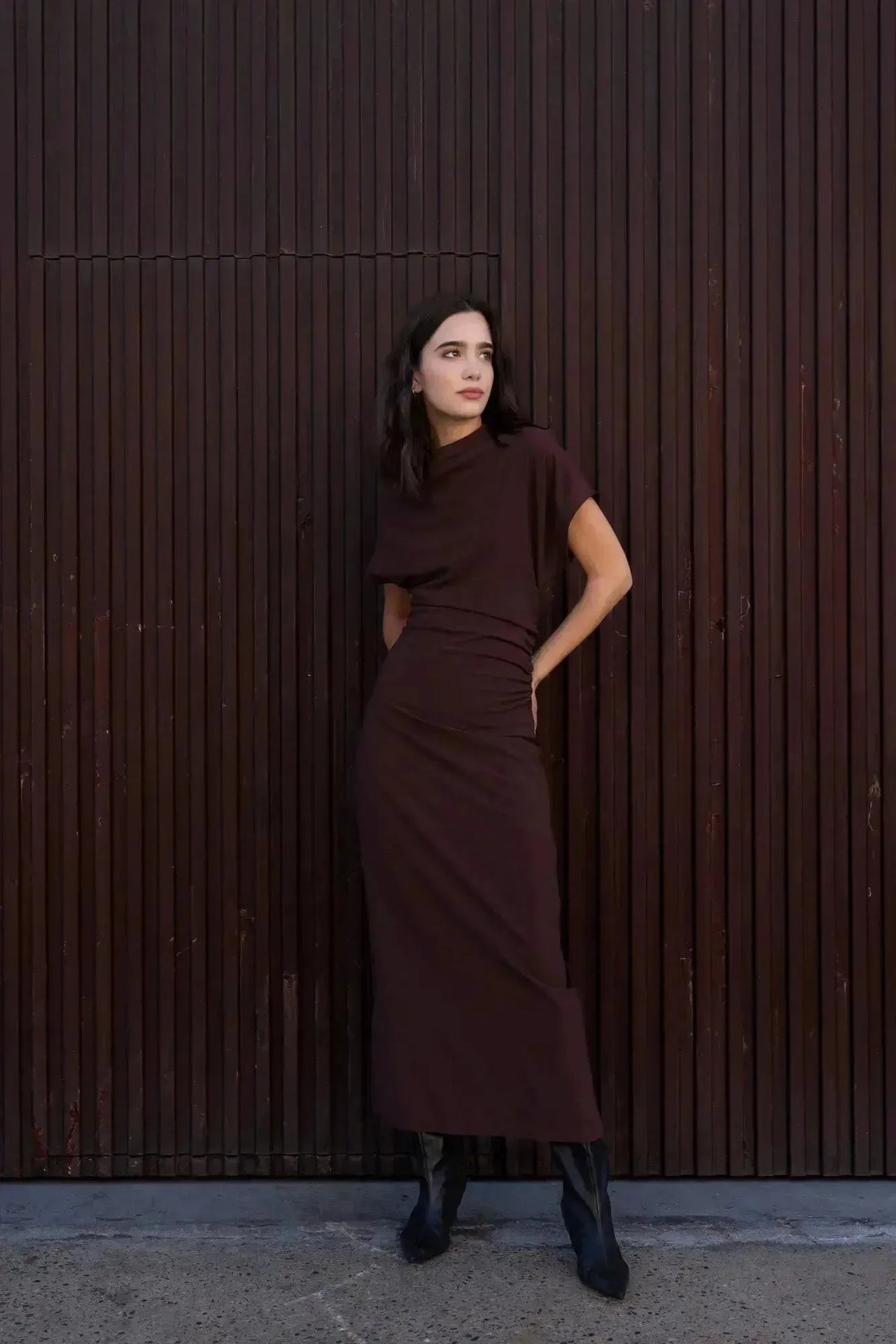 Robe longue marron ajustée femme avec détails drapés, modèle debout devant un mur en bois avec des bottes.