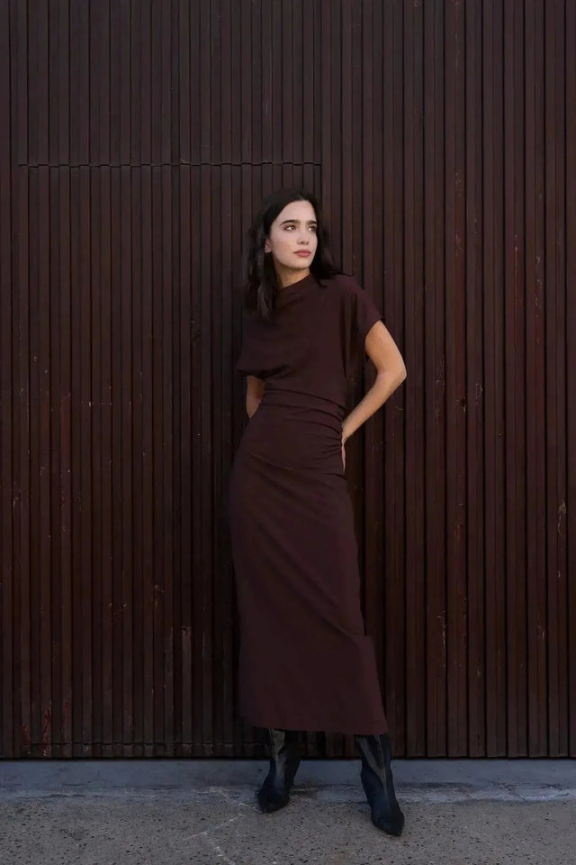 Robe longue marron ajustée femme avec détails drapés, modèle debout devant un mur en bois avec des bottes.