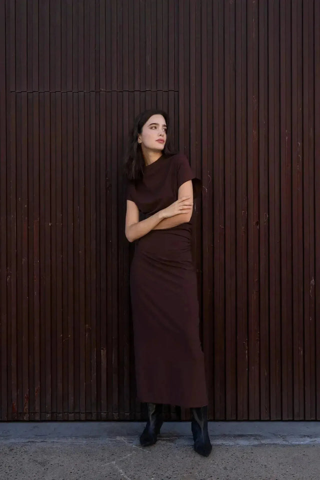 Robe longue marron ajustée femme avec détails drapés, modèle appuyée contre un mur en bois.