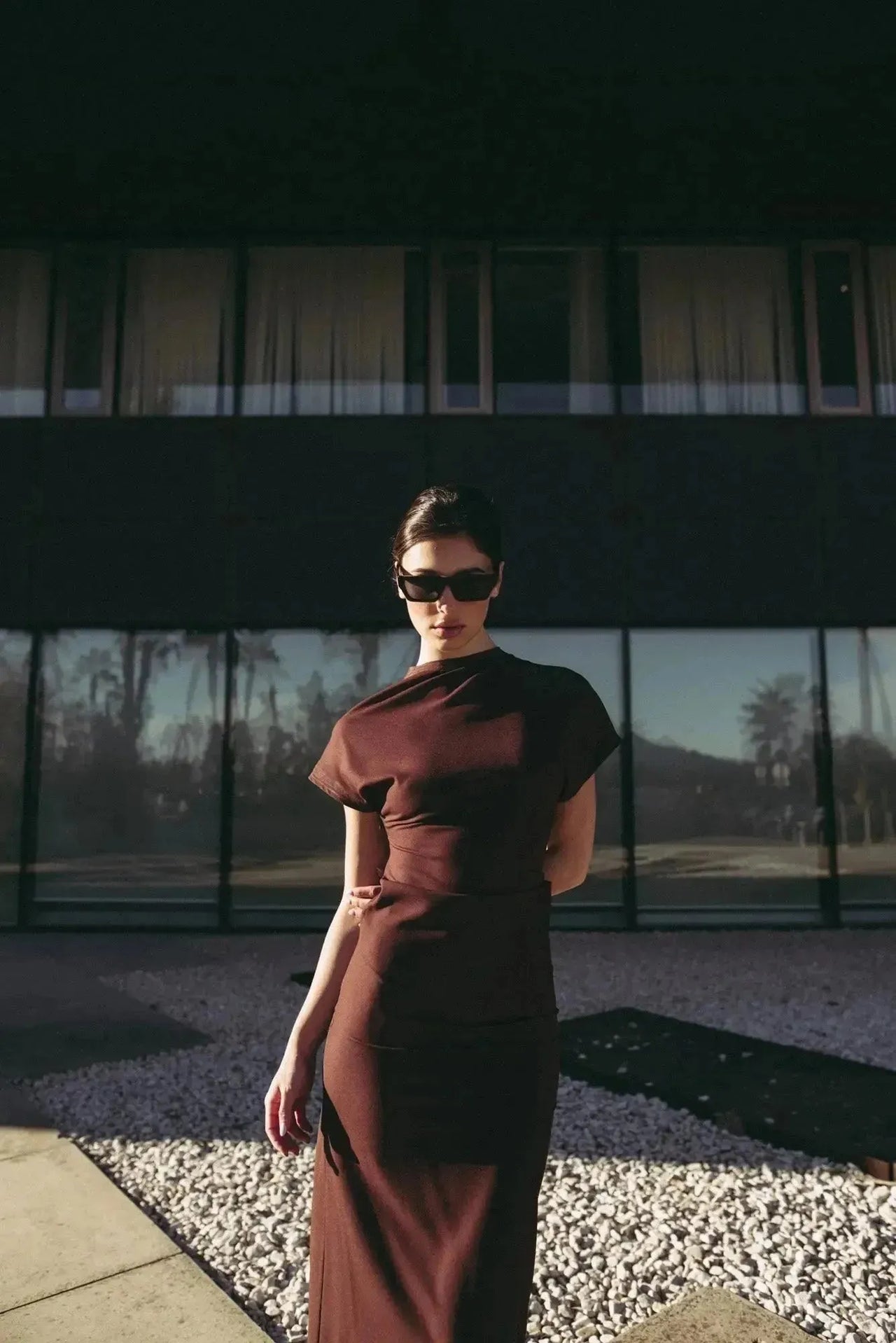 Robe longue marron ajustée femme avec détails drapés, modèle captivant regardant l'objectif en extérieur.