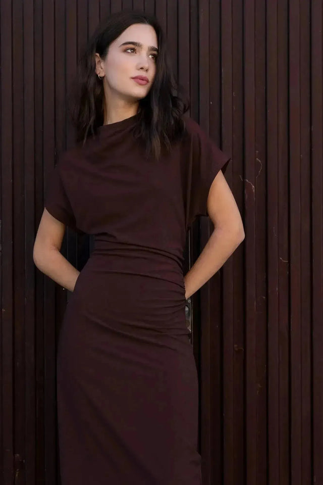 Robe longue marron ajustée femme avec détails drapés, modèle posant devant un mur en bois.