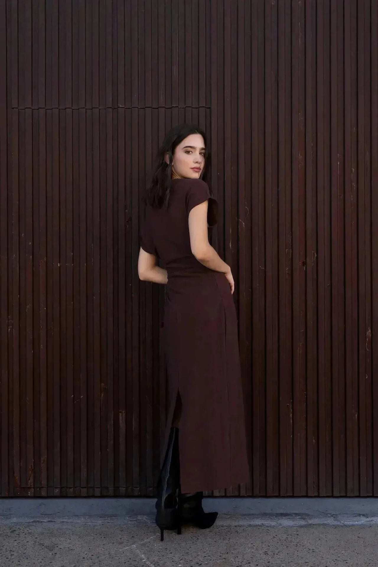 Robe longue marron ajustée femme avec détails drapés, vue de dos du modèle devant un mur en bois.