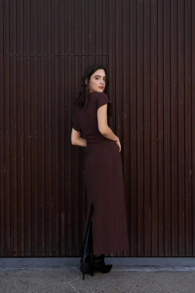Robe longue marron ajustée femme avec détails drapés, vue de dos du modèle devant un mur en bois.