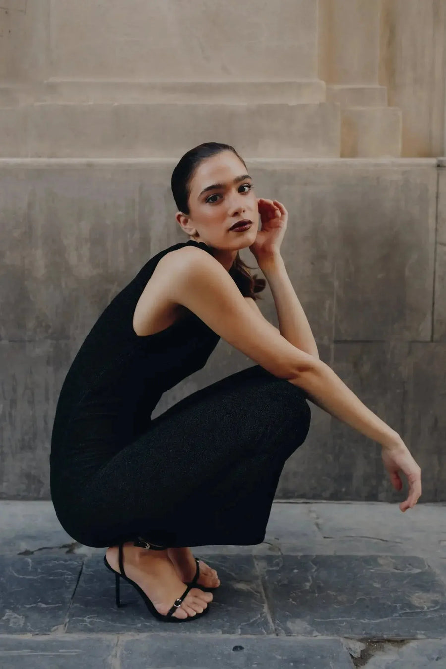 Femme assise dans une robe longue noire ajustée sans manches avec nœud, montrant le design moderne et chic, dans un cadre contemporain.
