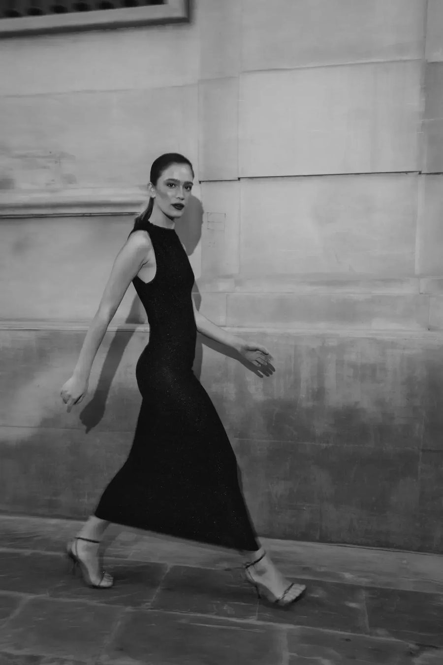 Femme marchant avec grâce dans une robe longue noire ajustée sans manches avec nœud, illustrant le mouvement et l'élégance de la pièce.
