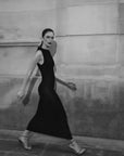 Femme marchant avec grâce dans une robe longue noire ajustée sans manches avec nœud, illustrant le mouvement et l'élégance de la pièce.