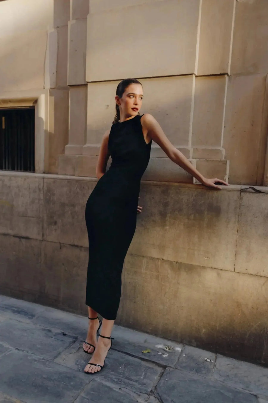 Modèle posant élégamment en robe longue noire ajustée sans manches avec nœud, mettant en avant son style minimaliste dans une ambiance urbaine.