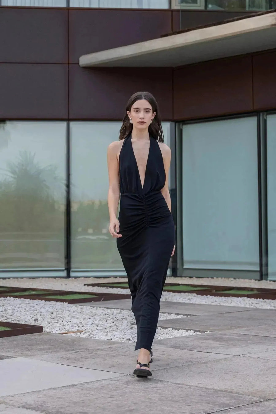 Modèle marchant dans une robe longue noire dos nu, affichant son look sophistiqué. Idéale pour les mariages, cette robe longue pour mariage invité chic attire tous les regards.