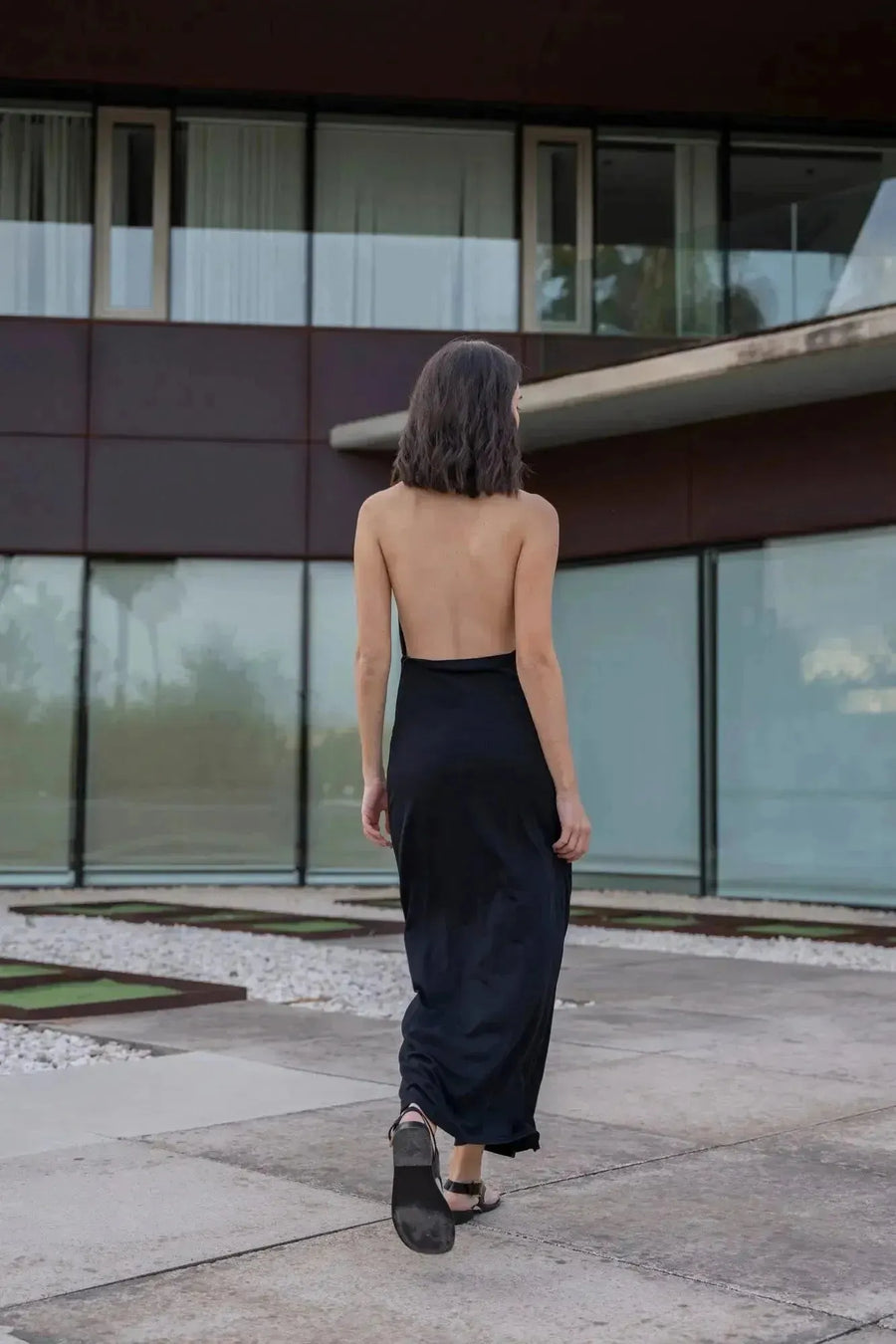 Vue arrière d'un modèle portant la robe longue noire dos nu, accentuant son design élégant. Une robe longue pour mariage invité chic pour un style inoubliable.