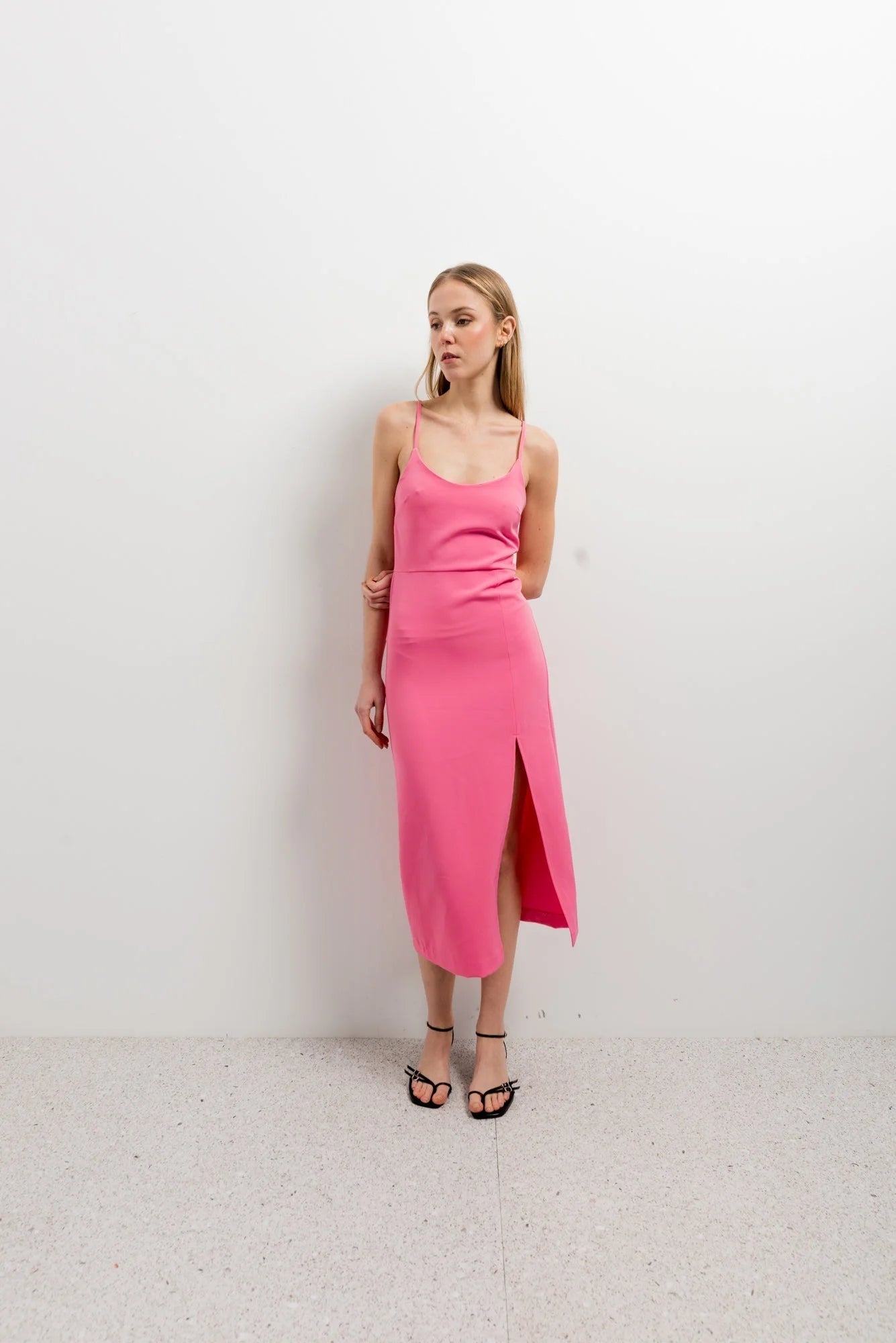 Modèle portant une robe rose midi à bretelles avec fente, élégante, confortable et ajustable, devant un mur blanc.
