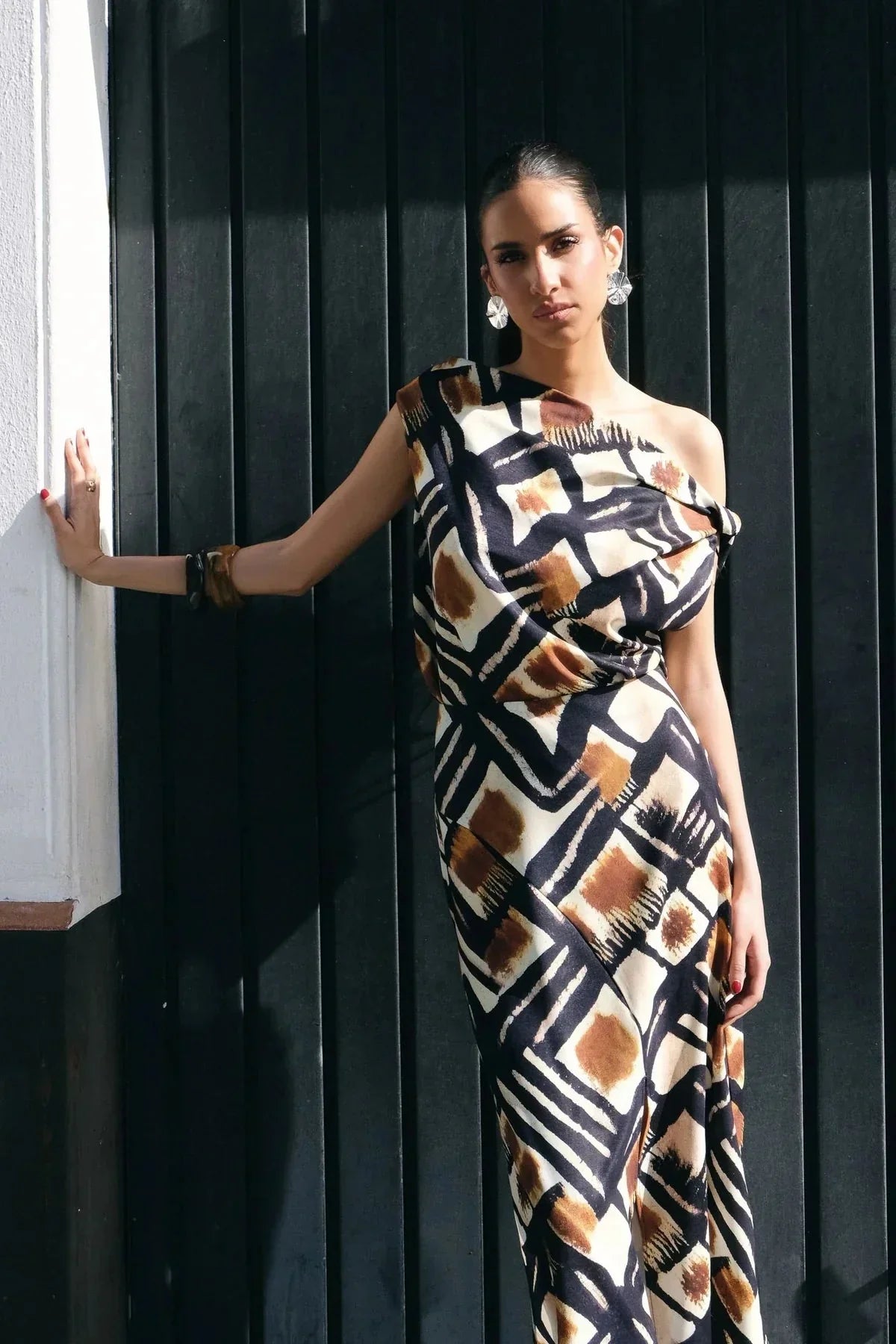 Femme en jupe longue motif graphique, affichant une pose élégante contre un mur. La jupe longue motif marron, noir et blanc souligne le style audacieux et moderne de cette pièce.