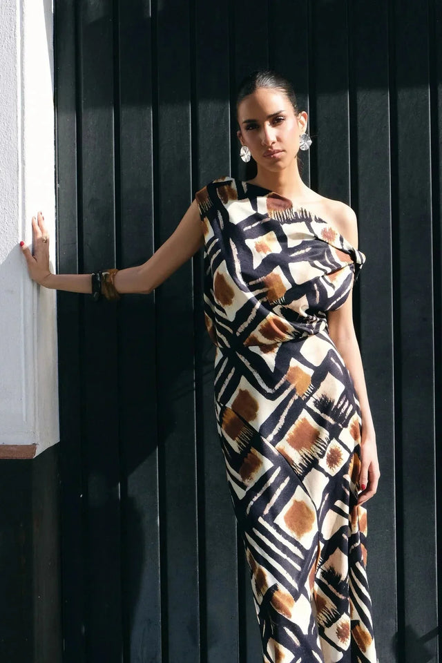 Femme en jupe longue motif graphique, affichant une pose élégante contre un mur. La jupe longue motif marron, noir et blanc souligne le style audacieux et moderne de cette pièce.