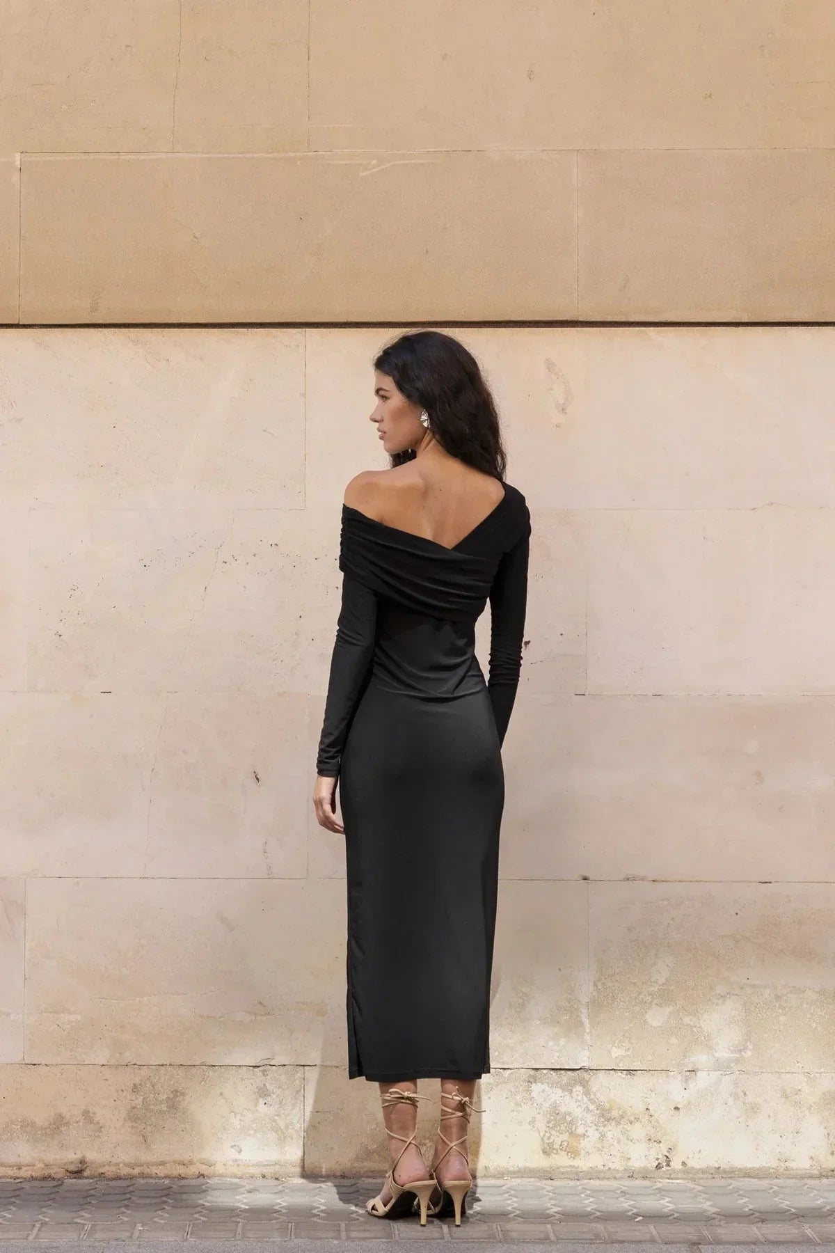 Femme en robe asymétrique noire longue à manches longues avec neckline drapé et talons beige