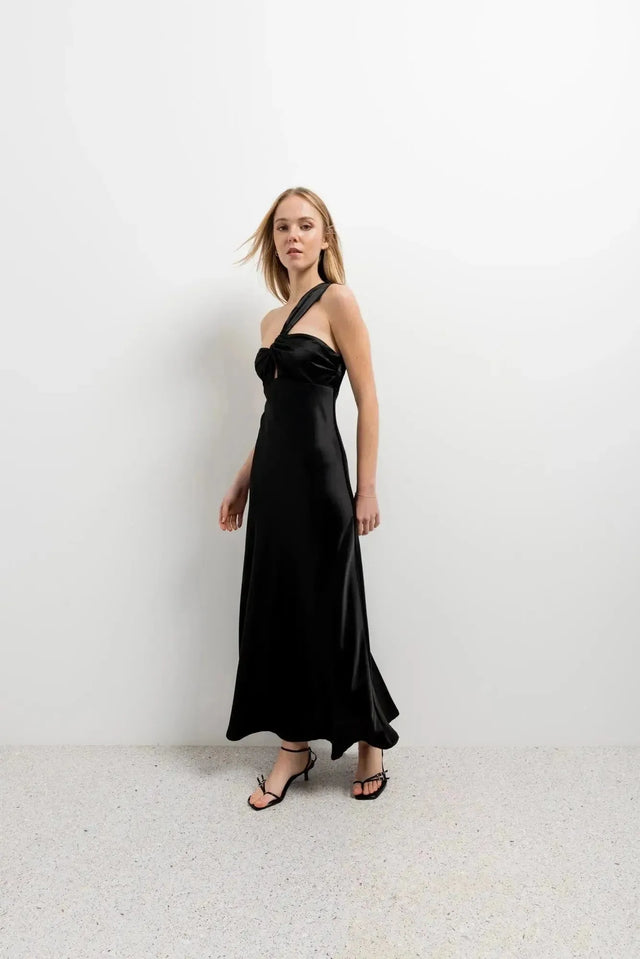 Modèle marchant dans une robe de soirée femme midi asymétrique noire, design unique avec encolure sublimante, parfaite pour les occasions spéciales.