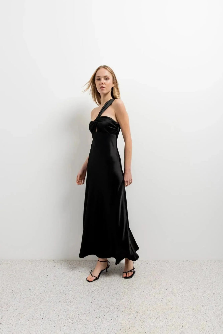 Modèle marchant dans une robe de soirée femme midi asymétrique noire, design unique avec encolure sublimante, parfaite pour les occasions spéciales.
