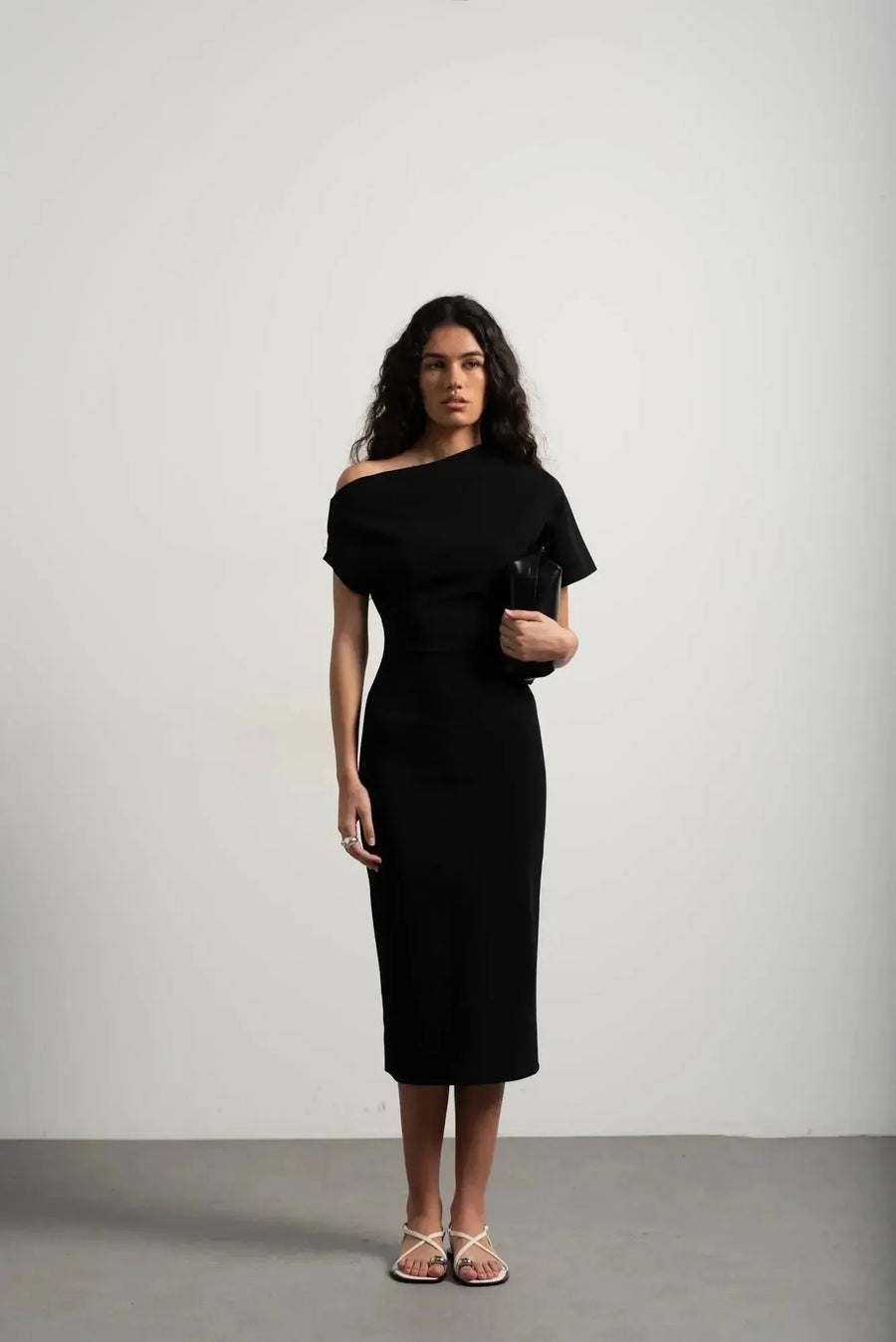 Robe asymétrique mi longue noire femme, élégance versatile pour soirées tendance 2024. Modèle féminin avec un décolleté asymétrique, porté avec une pochette noire et des sandales.