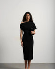 Robe asymétrique mi longue noire femme, élégance versatile pour soirées tendance 2024. Modèle féminin avec un décolleté asymétrique, porté avec une pochette noire et des sandales.