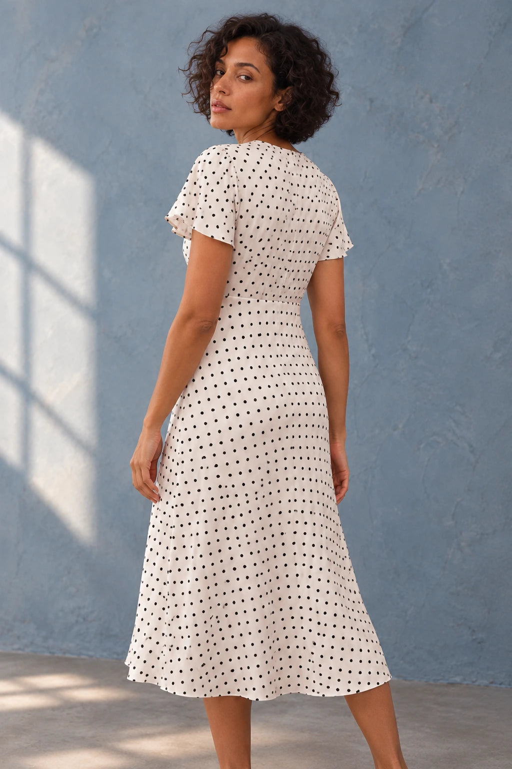 Femme vue de dos dans la robe à petit pois blancs et noirs, midi fluide, décolleté V, manches volantées chic, affichant un style sophistiqué.