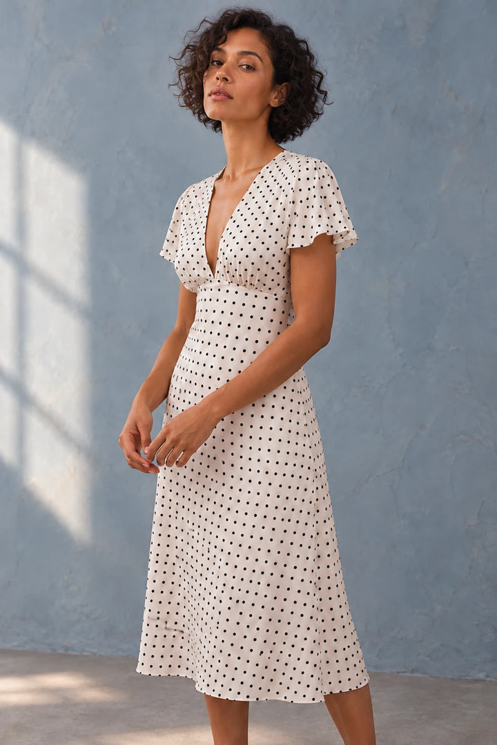 Modèle féminine portant la robe à petit pois blancs et noirs, midi fluide, décolleté V, manches volantées chic, se tenant en pose élégante.