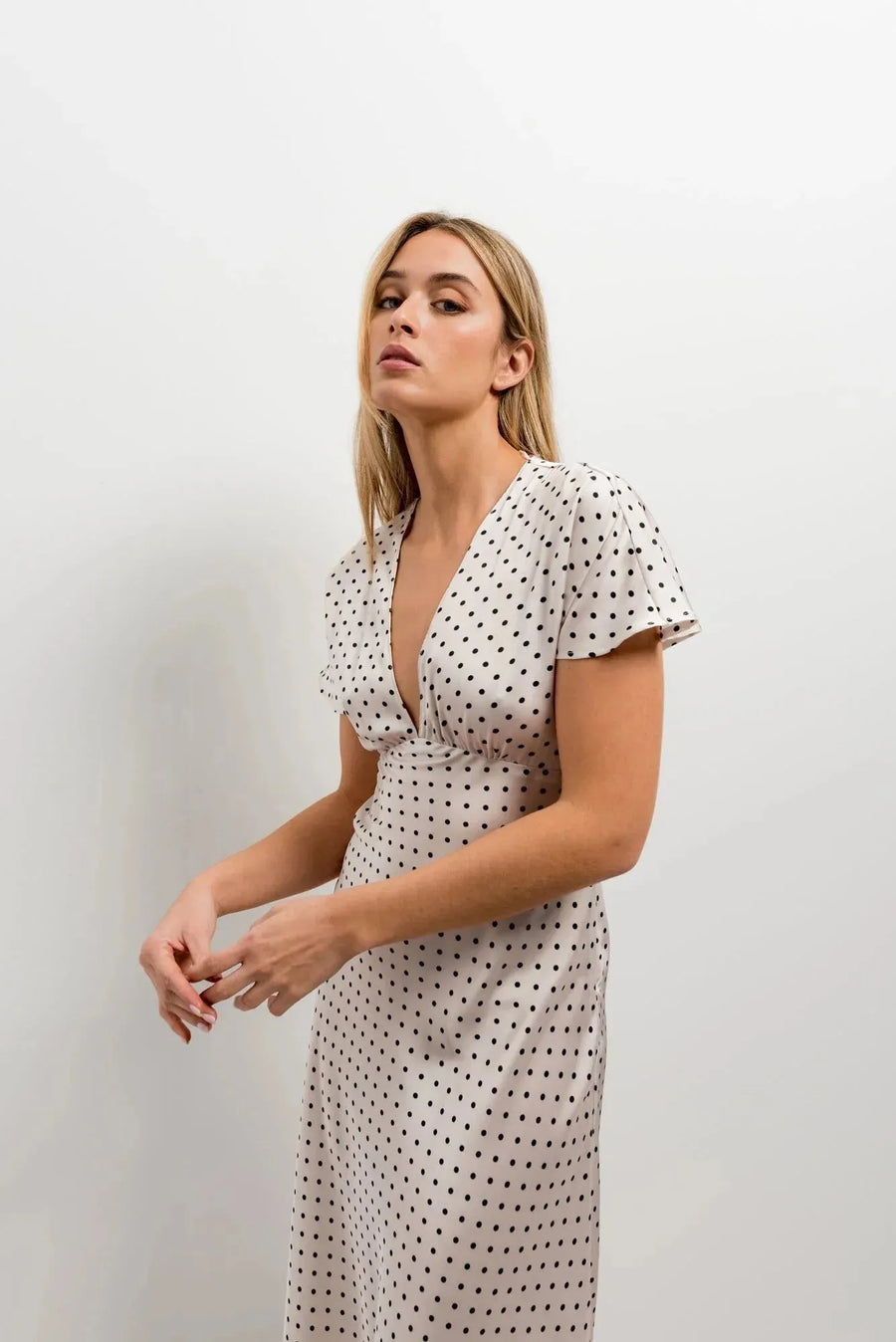 Modèle féminine portant la robe à petit pois blancs et noirs, midi fluide, décolleté V, manches volantées chic, se tenant en pose élégante.