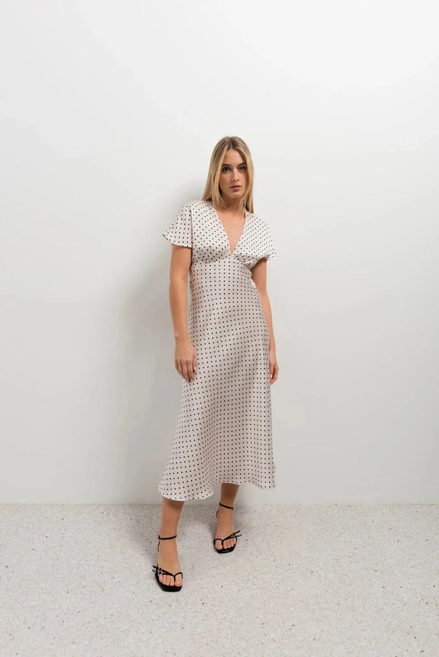 Femme posant avec assurance dans la robe à petit pois blancs et noirs, midi fluide, décolleté V, manches volantées chic, sur fond minimaliste.
