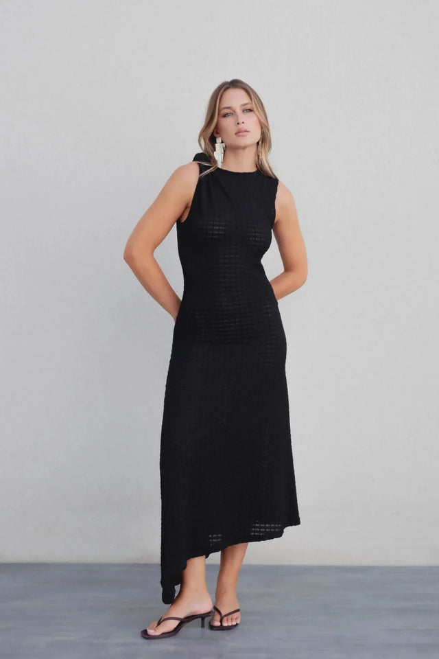 Femme posant dans une élégante robe maille noire asymétrique moulante avec détail noué à une épaule – longueur midi, affichant un look moderne et décontracté.
