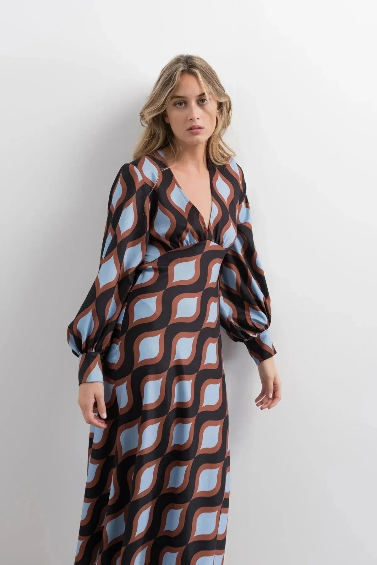 Femme debout dans une robe motif géométrique midi à décolleté V profond et manches bouffantes, avec des couleurs marron et bleu clair qui captent le regard.