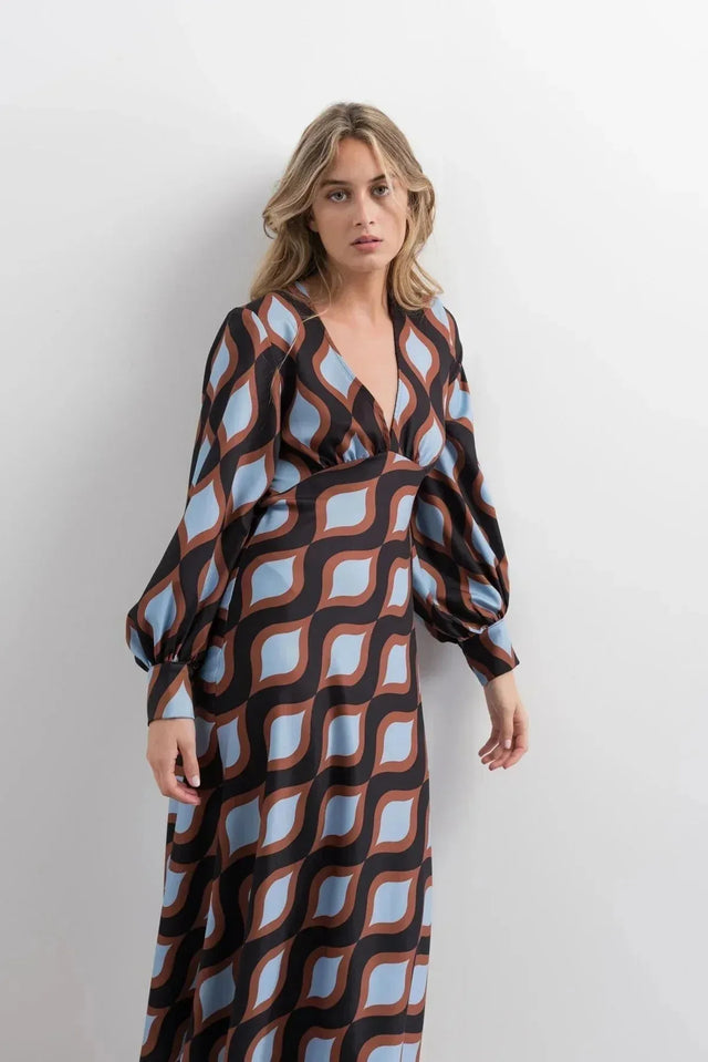 Femme debout dans une robe motif géométrique midi à décolleté V profond et manches bouffantes, avec des couleurs marron et bleu clair qui captent le regard.
