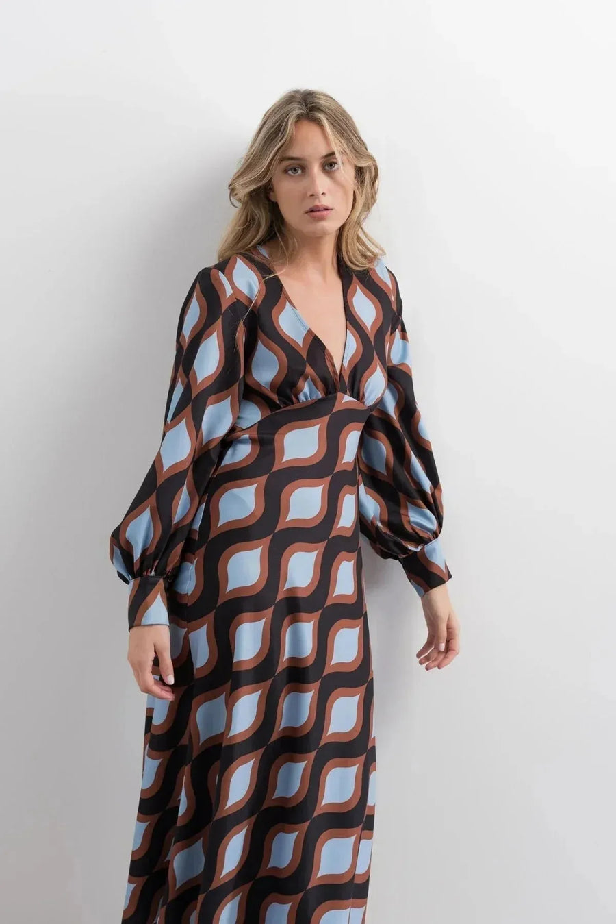 Femme debout dans une robe motif géométrique midi à décolleté V profond et manches bouffantes, avec des couleurs marron et bleu clair qui captent le regard.