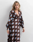 Femme debout dans une robe motif géométrique midi à décolleté V profond et manches bouffantes, avec des couleurs marron et bleu clair qui captent le regard.