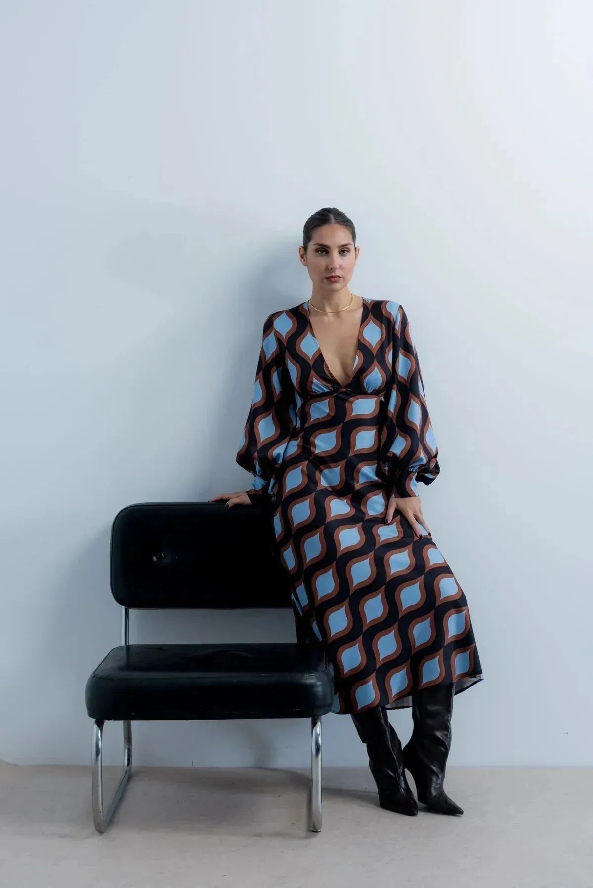 Modèle portant une robe motif géométrique midi au décolleté V et manches bouffantes, assise sur une chaise moderne.