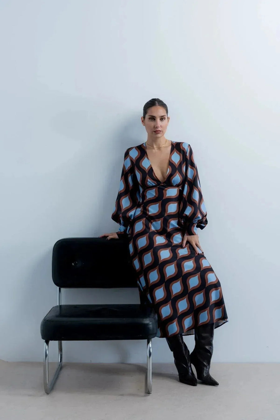 Modèle portant une robe motif géométrique midi au décolleté V et manches bouffantes, assise sur une chaise moderne.