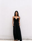 Femme en robe midi noire à fines bretelles, se tenant debout contre un mur, un look décontracté d'été. Une belle option de robe noire longue dos nu.