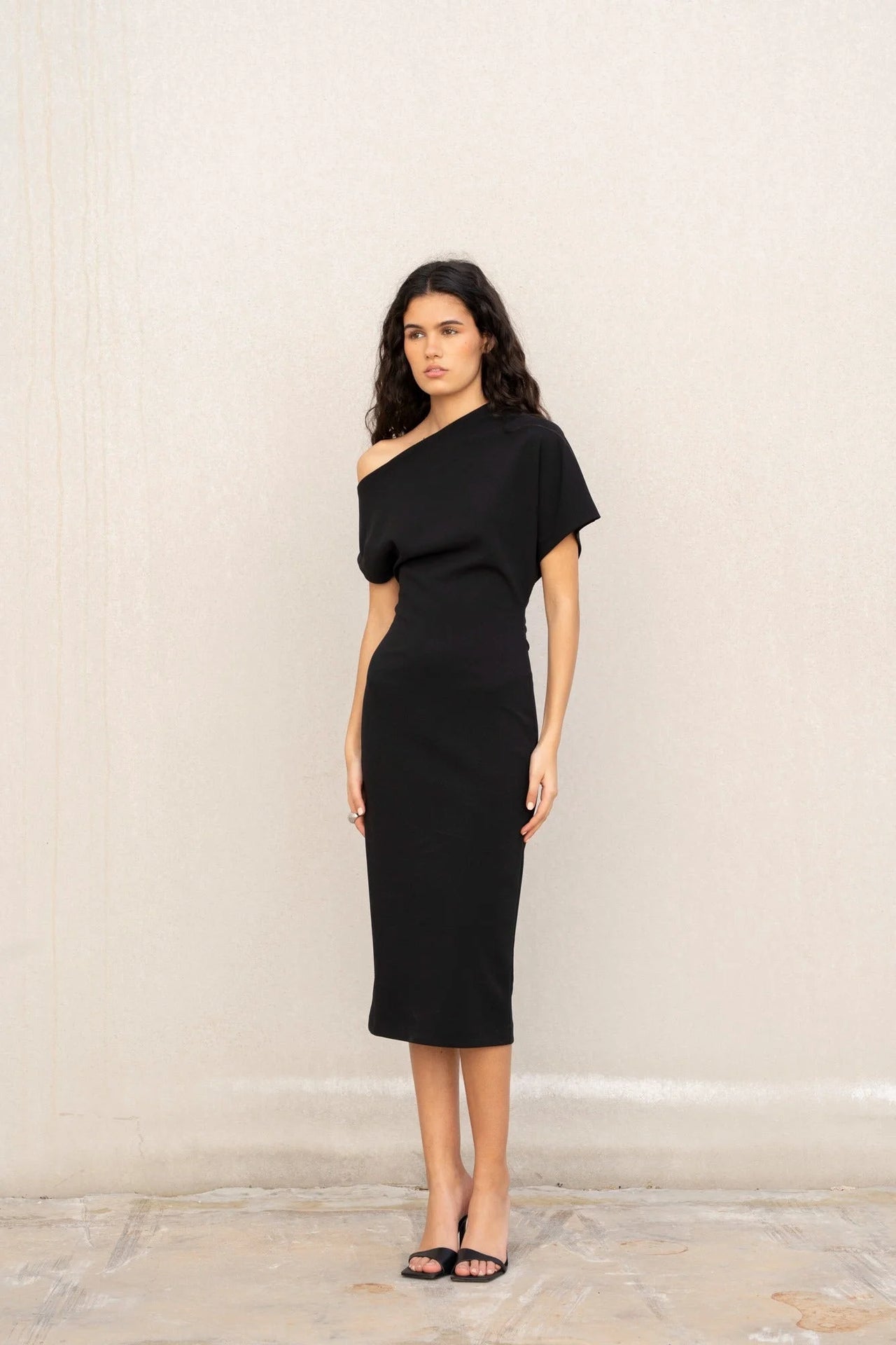 Modèle dans une robe asymétrique noire à épaule dénudée chic moulante midi pour soirée élégante, portant des sandales à talons.