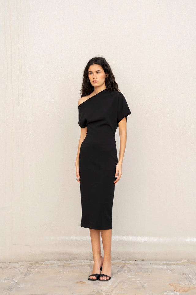 Modèle dans une robe asymétrique noire à épaule dénudée chic moulante midi pour soirée élégante, portant des sandales à talons.