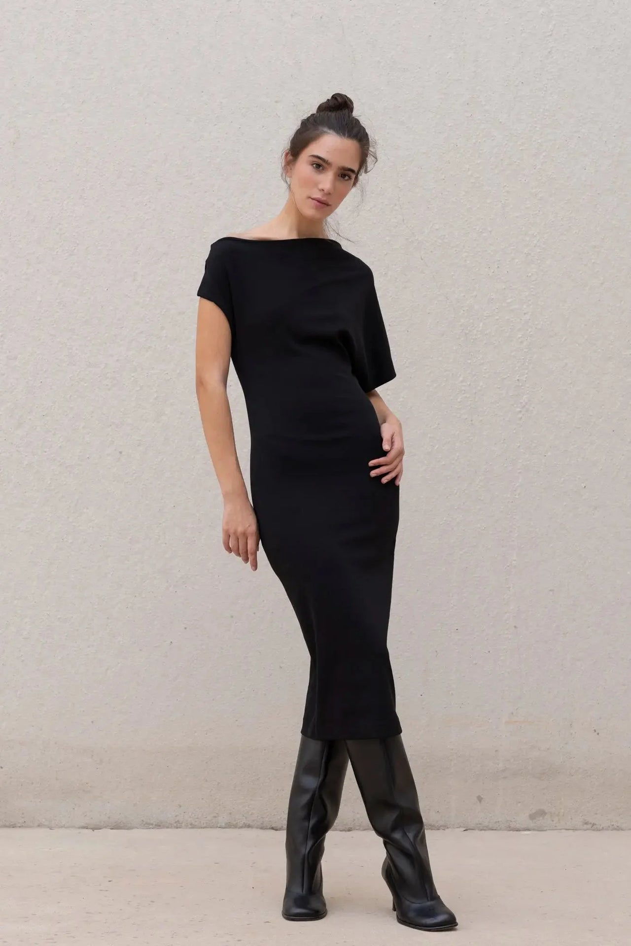 Femme en pose élégante dans une robe asymétrique noire à épaule dénudée chic moulante midi pour soirée, avec des bottes noires.