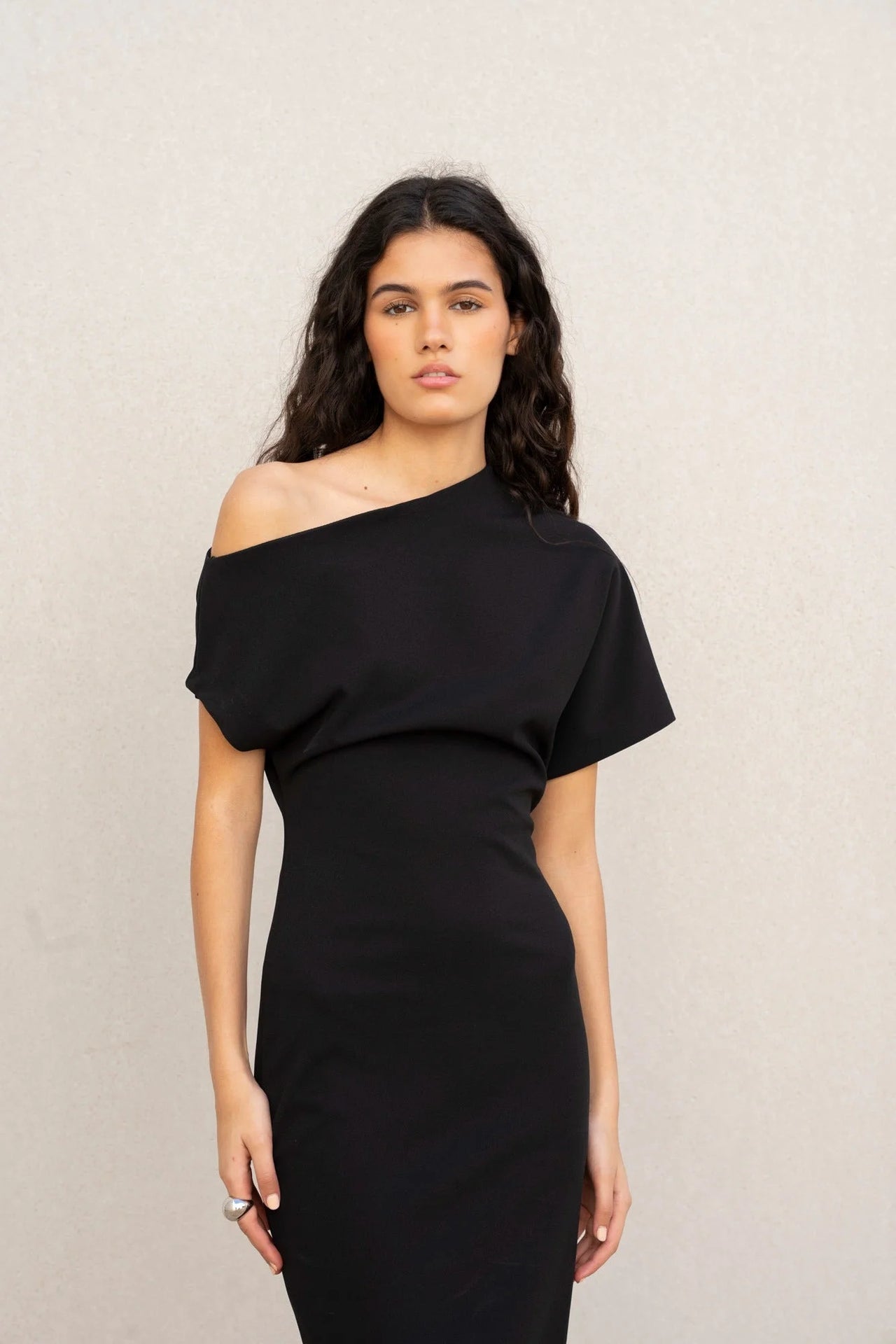 Vue frontale d'un modèle portant une robe asymétrique noire à épaule dénudée chic moulante midi pour soirée élégante, avec un regard captivant.
