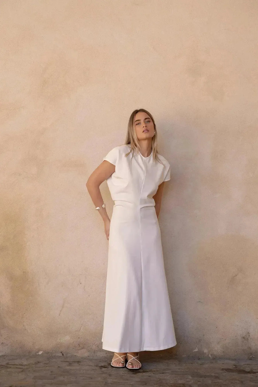 Femme en robe longue blanche chic, profilée avec un design plissé, se tenant debout dans un décor sobre.