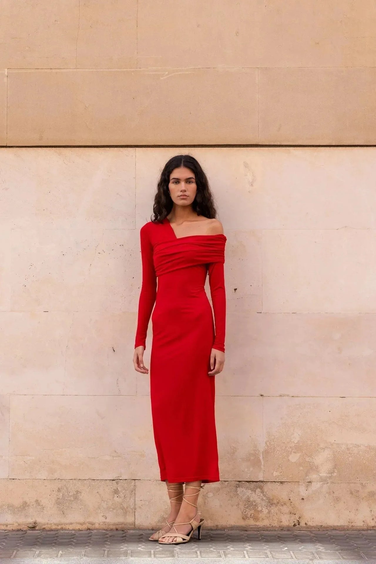 Robe Rouge Moulante Asymétrique à Épaules Découvertes | Modèle en rouge moulant devant un mur, parfaite pour vos soirées élégantes.