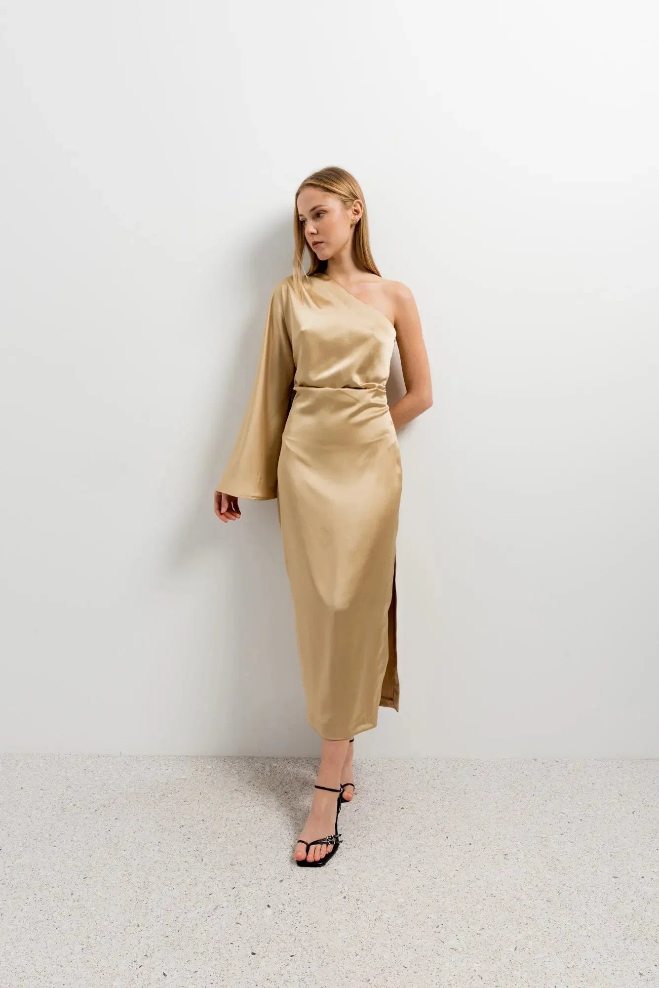 Modèle portant la Robe Dorée Satinée Asymétrique Une Manche, avec fente latérale, sur un fond sobre, parfaite pour les occasions spéciales.