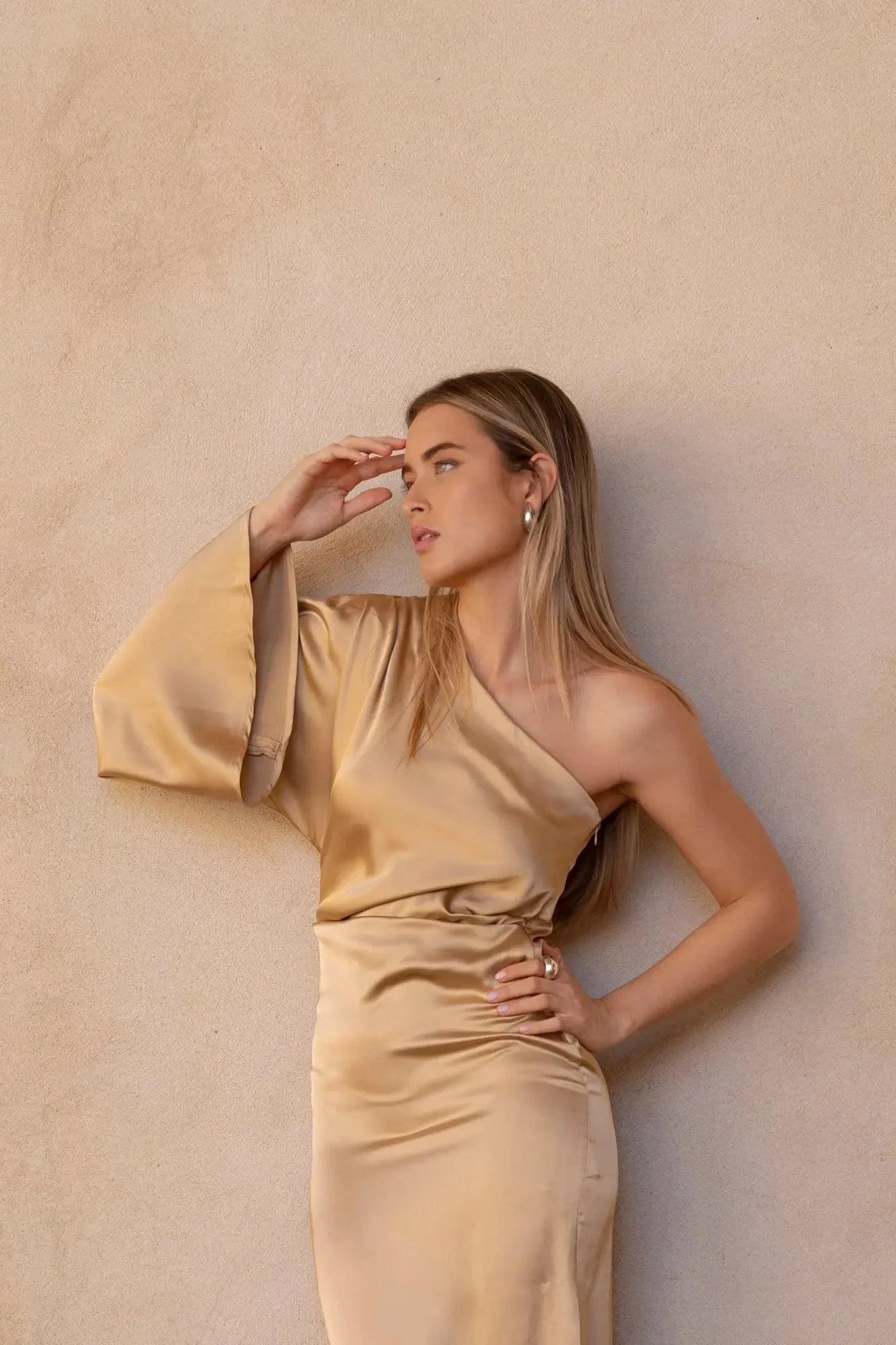Détail de la Robe Dorée Satinée Asymétrique Une Manche Avec Fente Latérale, montrant une femme en train de jouer avec ses cheveux, chic et stylée.