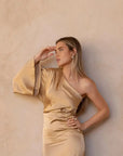 Détail de la Robe Dorée Satinée Asymétrique Une Manche Avec Fente Latérale, montrant une femme en train de jouer avec ses cheveux, chic et stylée.