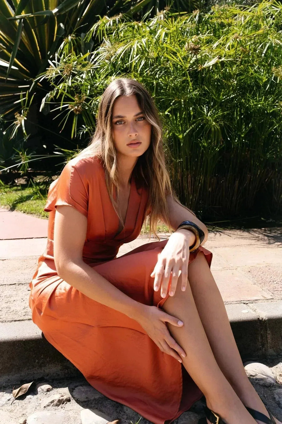 Femme assise dans un jardin, vêtue d'une robe midi satinée orange cuivré,款表现出te l'élégance naturelle de cette robe satin orange.