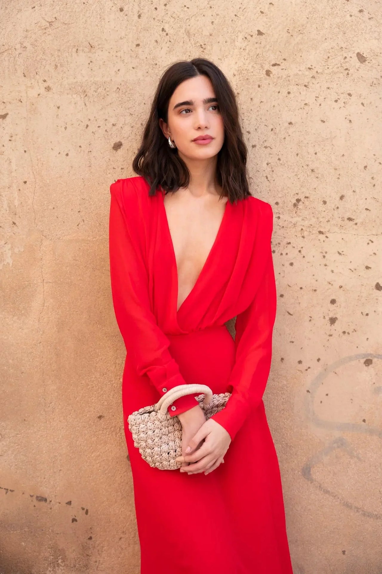 Femme portant une robe mousseline rouge midi à manches longues, décolleté plongeant élégant, accompagnée d'une pochette, parfaite pour des événements.
