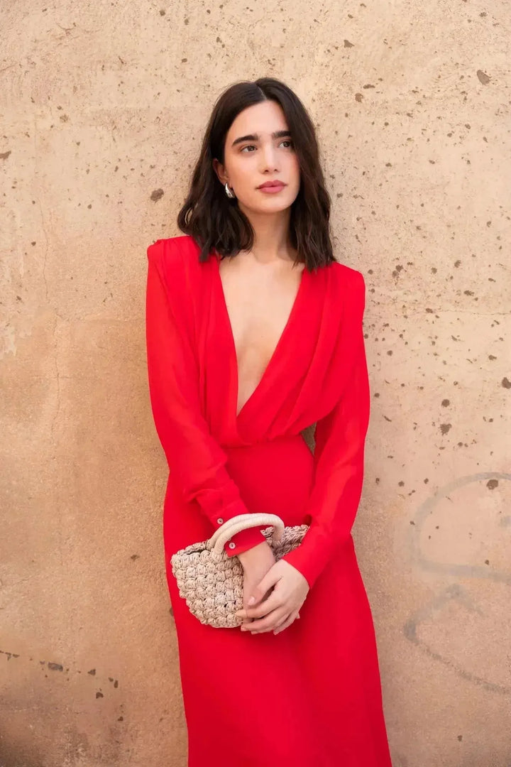 Femme portant une robe mousseline rouge midi à manches longues, décolleté plongeant élégant, accompagnée d'une pochette, parfaite pour des événements.