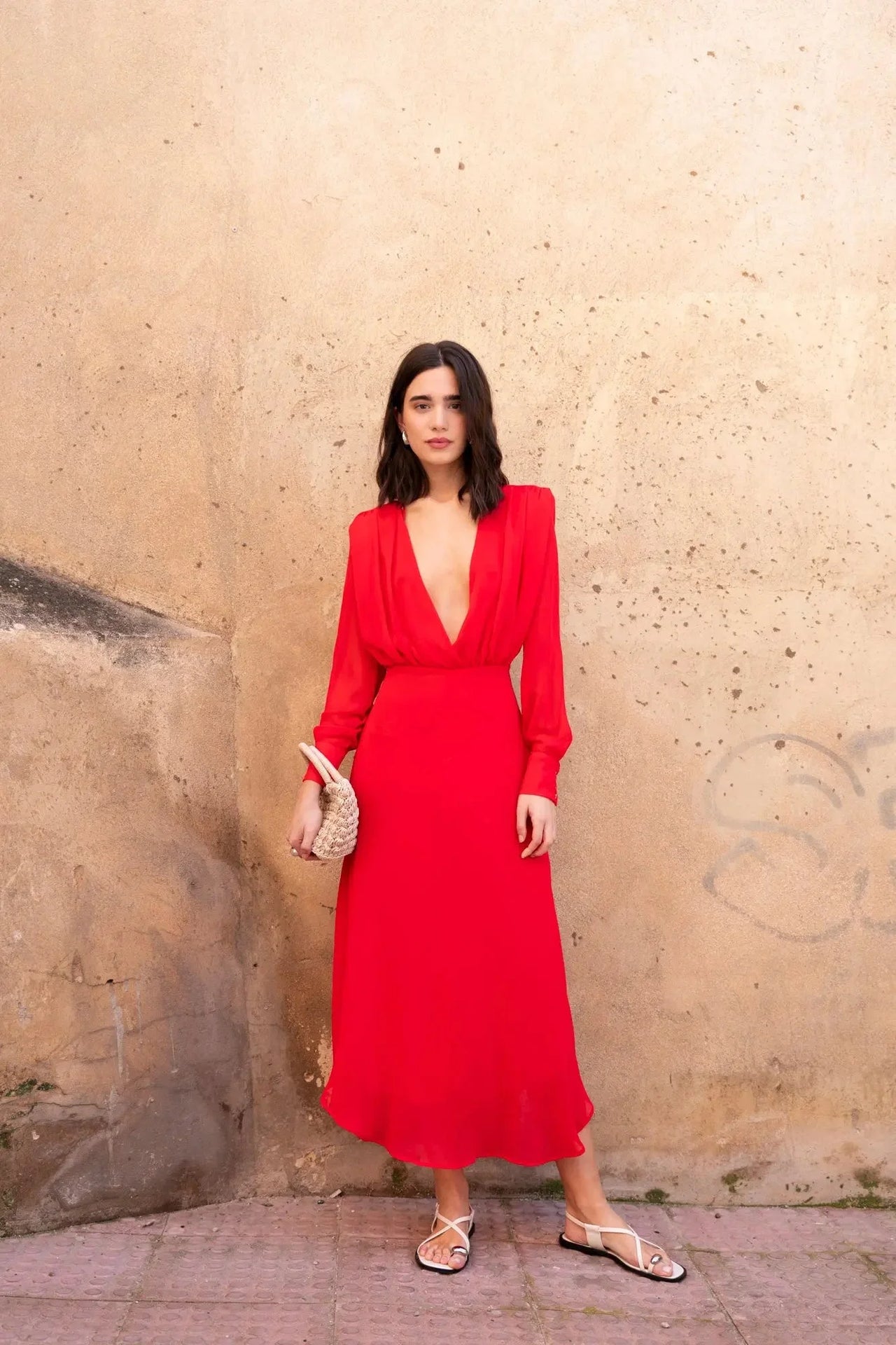 Robe mousseline rouge midi, décolleté plongeant, vue complète d'une femme posant avec une pochette, idéale pour un mariage ou une cérémonie.