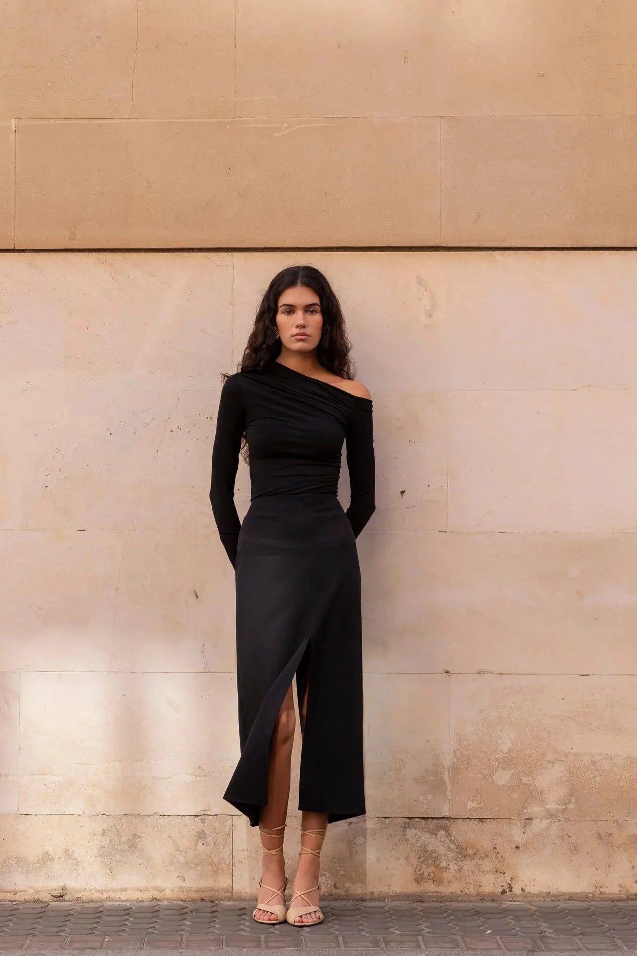 Modèle debout contre un mur avec une robe noire longue femme élégante, mettant en avant son style chic.