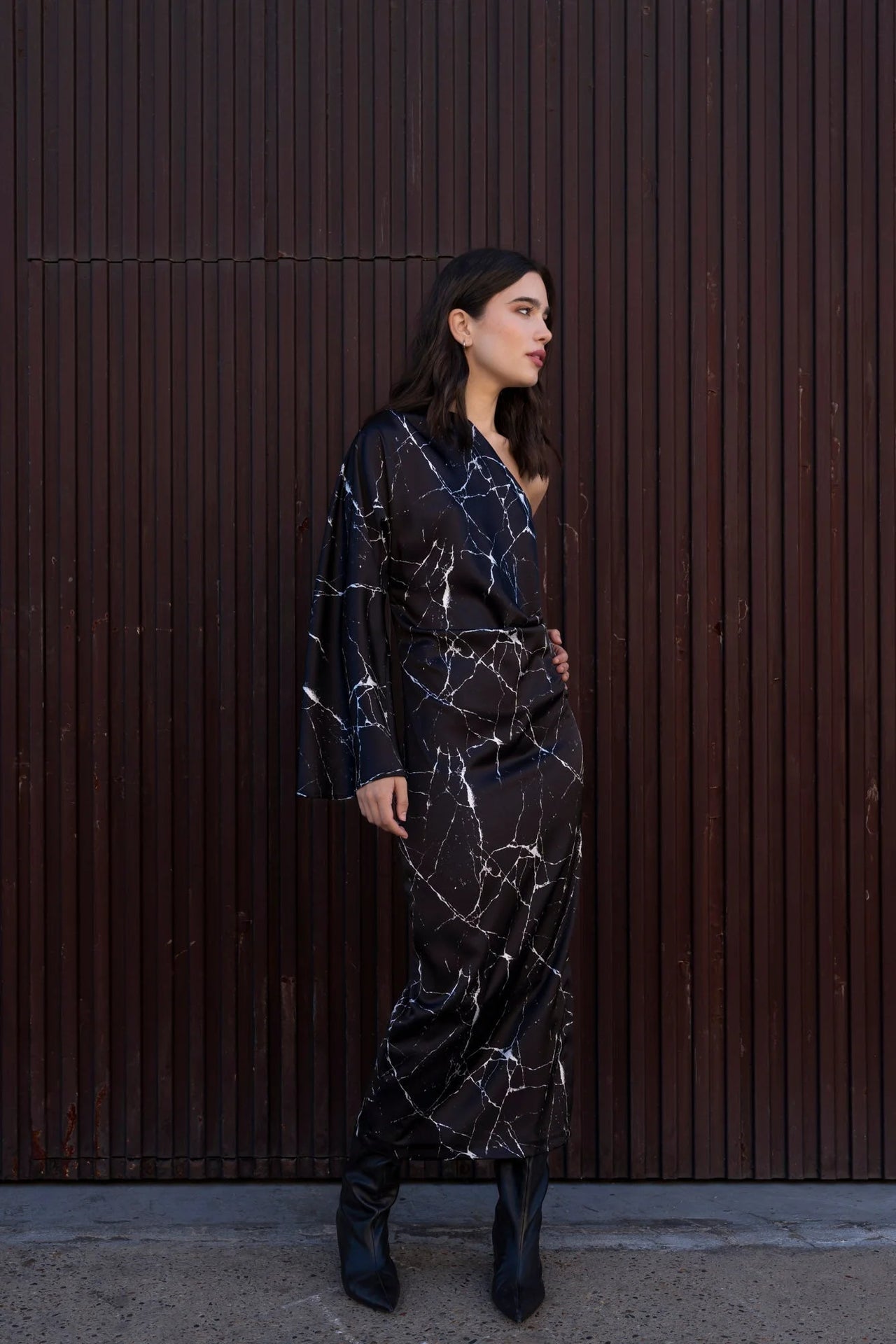 Femme portant la Robe de Soirée Femme Asymétrique Satinée Noire Avec Imprimé Marbre, vue en entier avec un fond en bois.